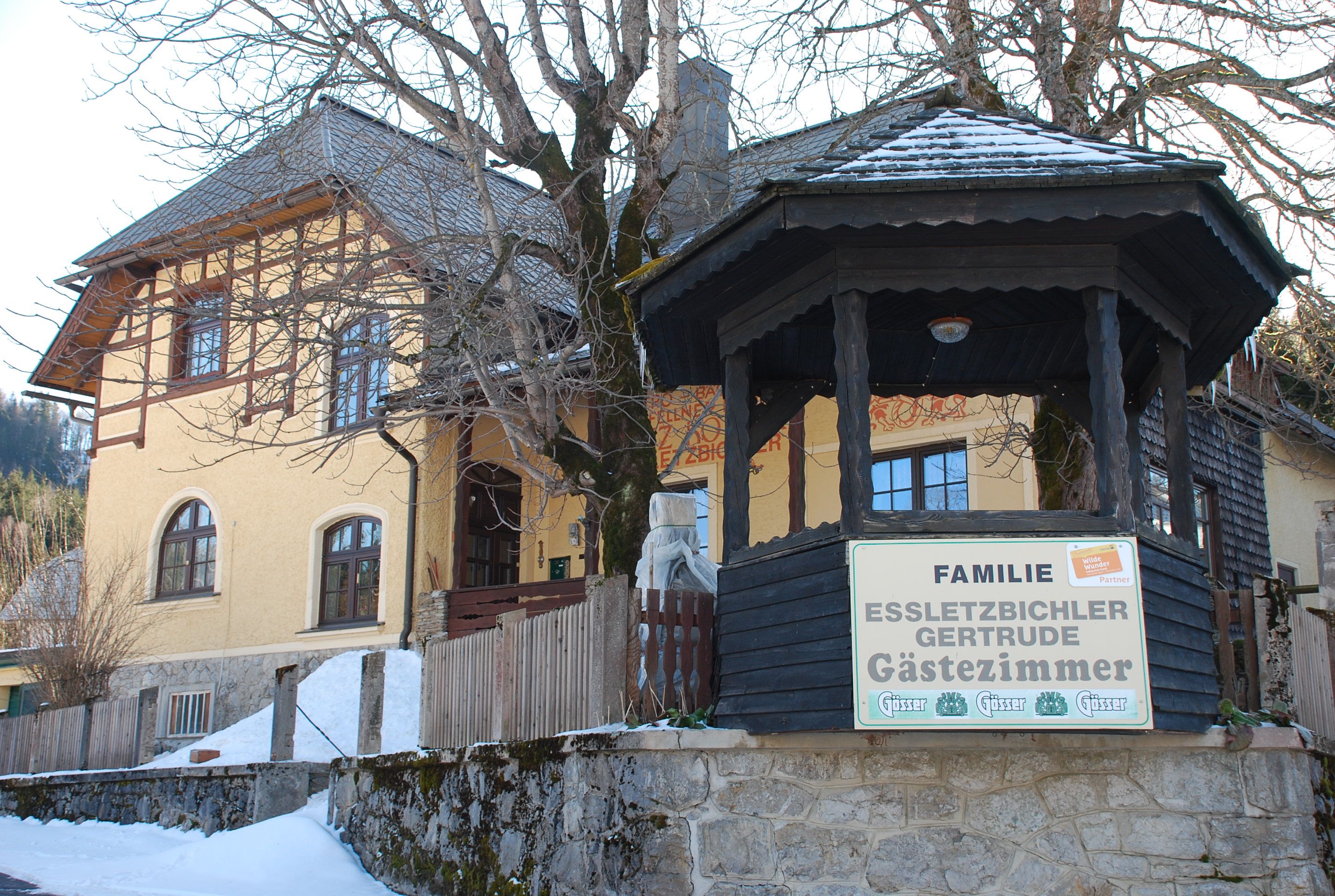 Žltý penzión s drevenou verandou a nápisom "Familie Essletzbichler Gertrude Gästezimmer".