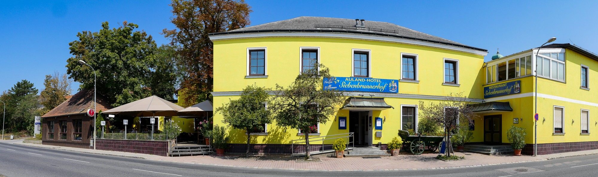Žltá budova s reštauráciou a hotelom, obklopená stromami a cestou.
