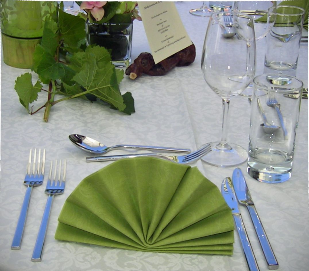 Elegantná dekorácia stola so zeleným skladaným obrúskom, príborom, pohárom na víno a pohárom na vodu na bielom obruse.