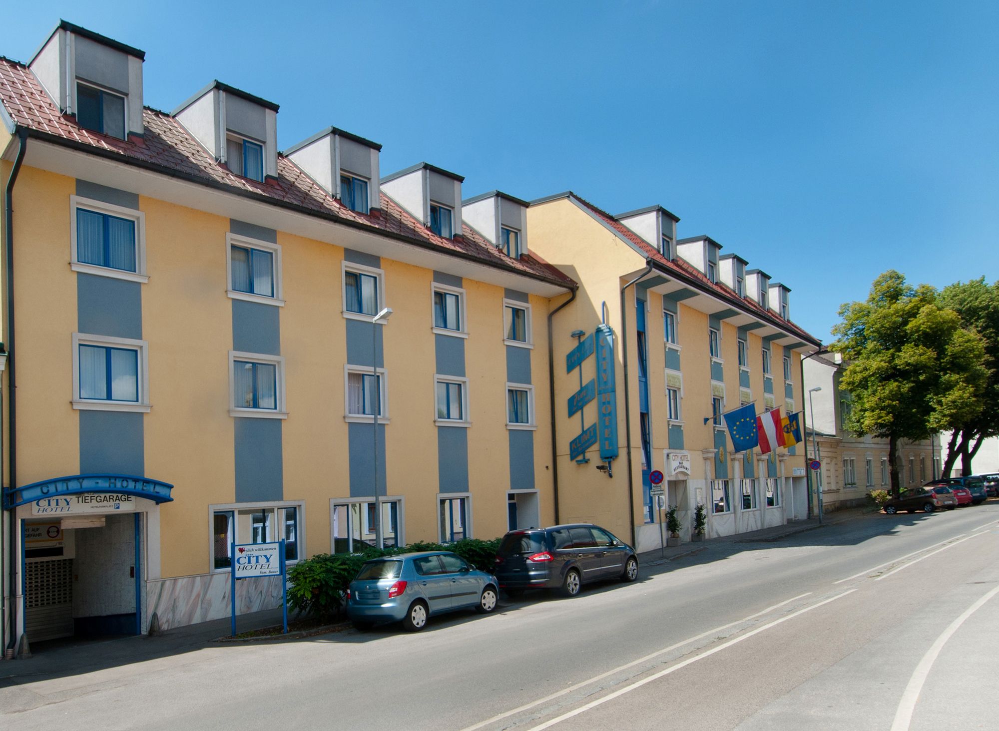 Exteriérový pohľad na žlto-modrý hotel s niekoľkými oknami a zaparkovanými autami pred ním.