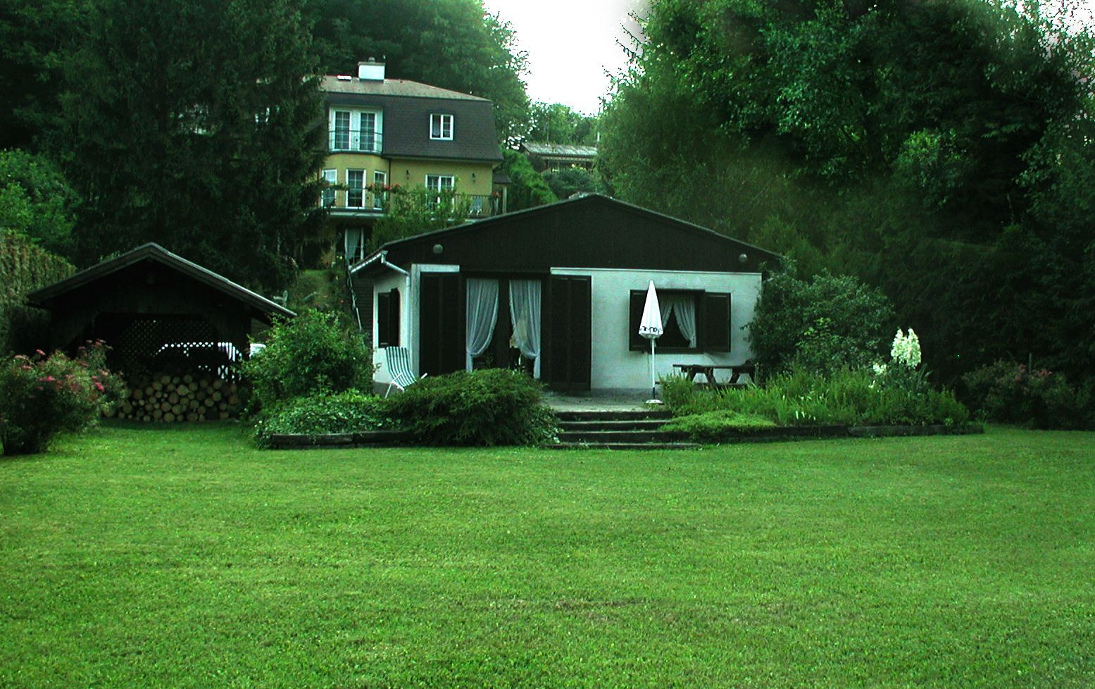 Malý domček so záhradou a terasou, obklopený stromami a väčšou budovou v pozadí.
