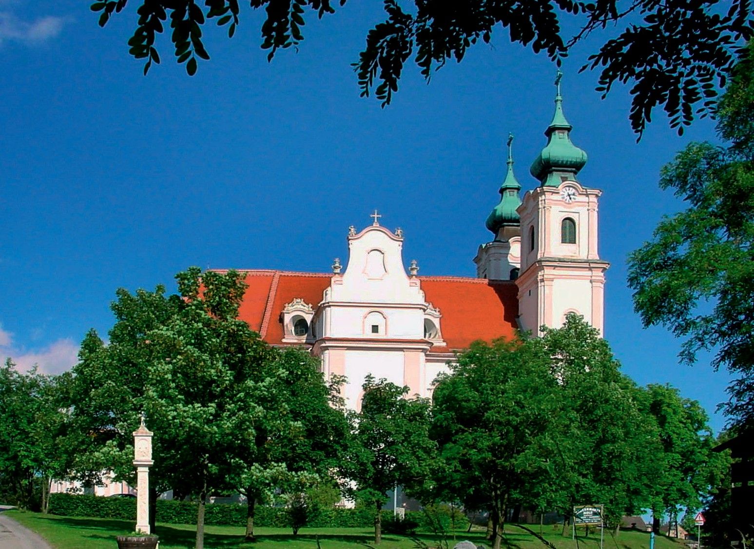 Bazilika Maria Dreieichen s červenými strechami a zelenými vežami, obklopená stromami.
