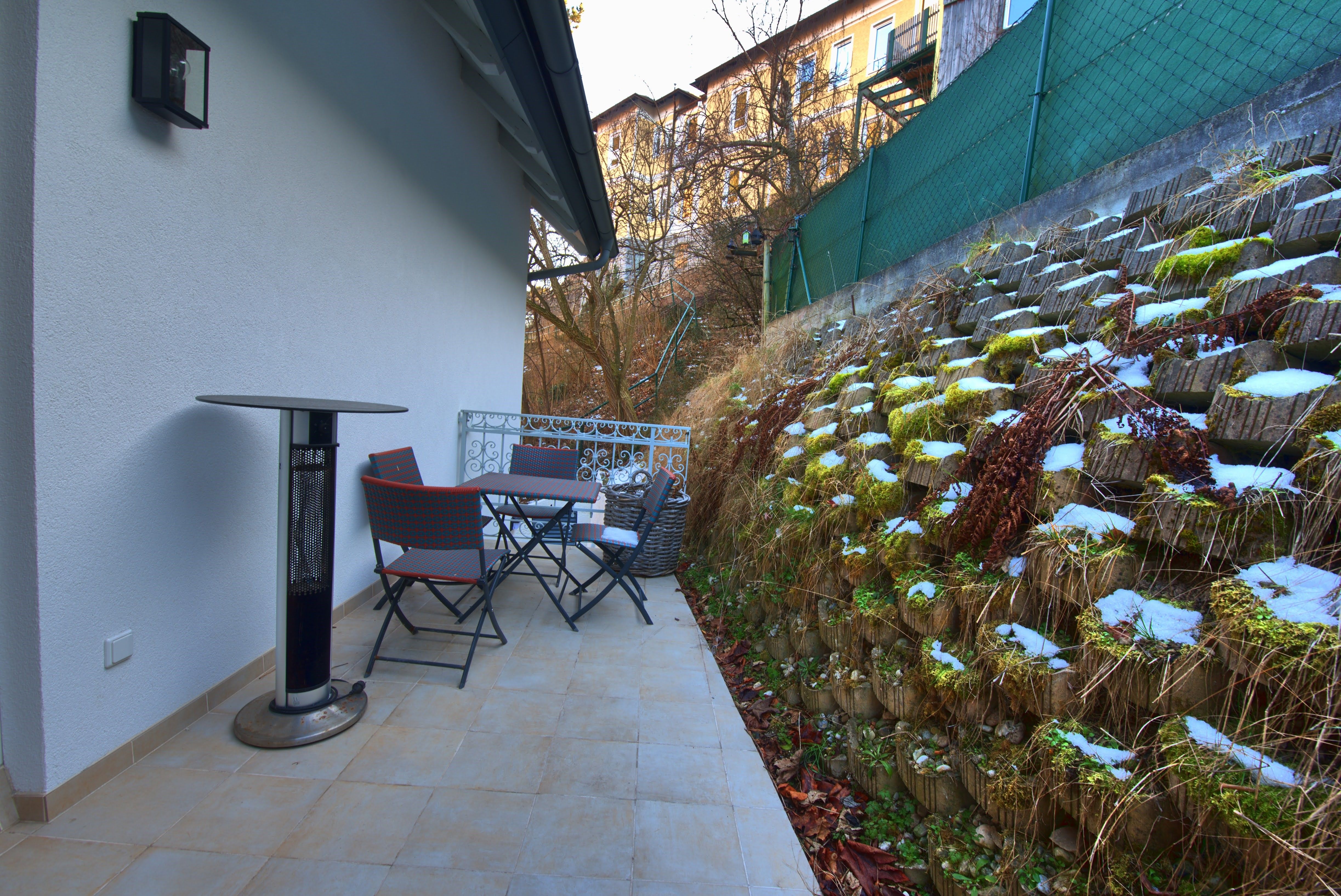 Terasa so stolom, stoličkami a sálavým vykurovacím telesom vedľa zelenej steny.