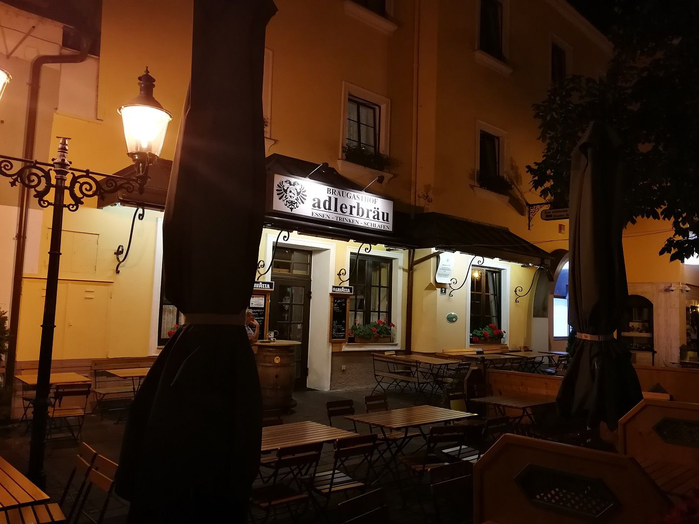 Nočný záber na reštauráciu s nápisom "Adlerbräu" a prázdnymi stolmi vonku.