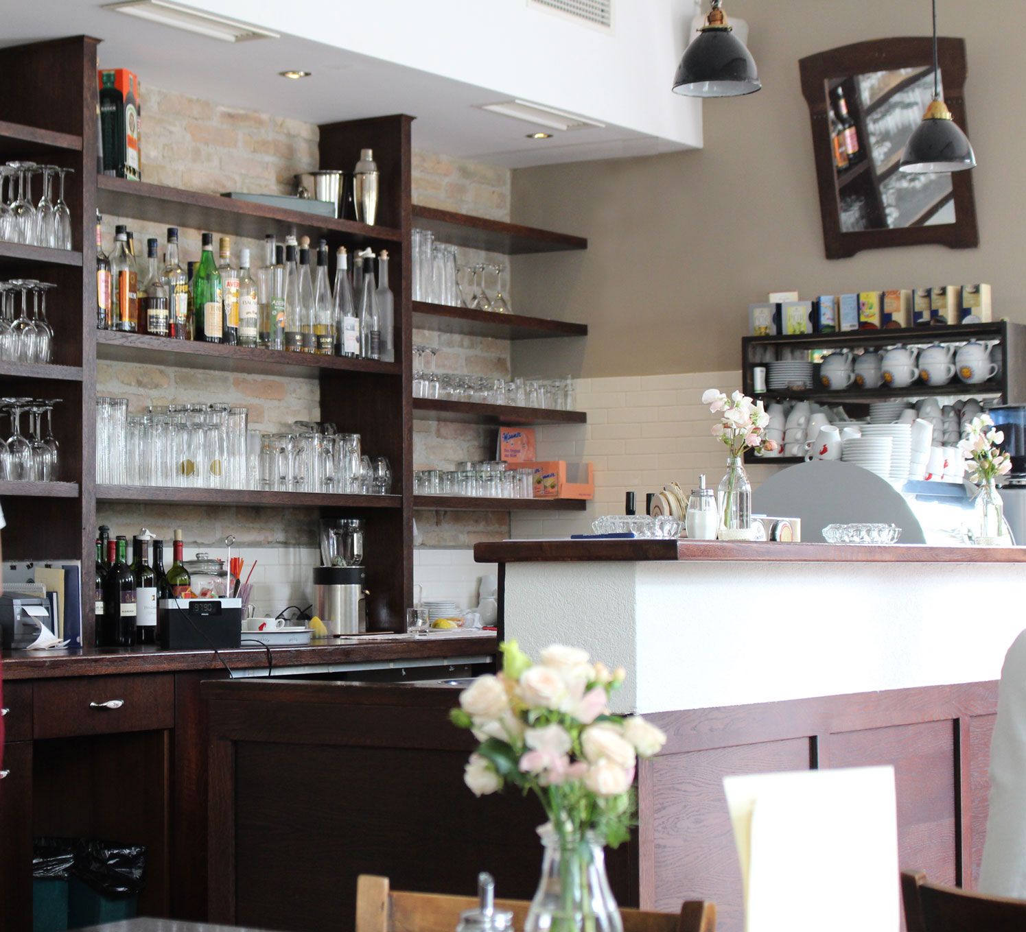 Pohľad do interiéru baru s policami plnými pohárov a fliaš, kvety na pulte.