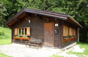 Malý rustikálny drevený poľovnícky zámoček s okenicami a parožím na fasáde, obklopený lesom.