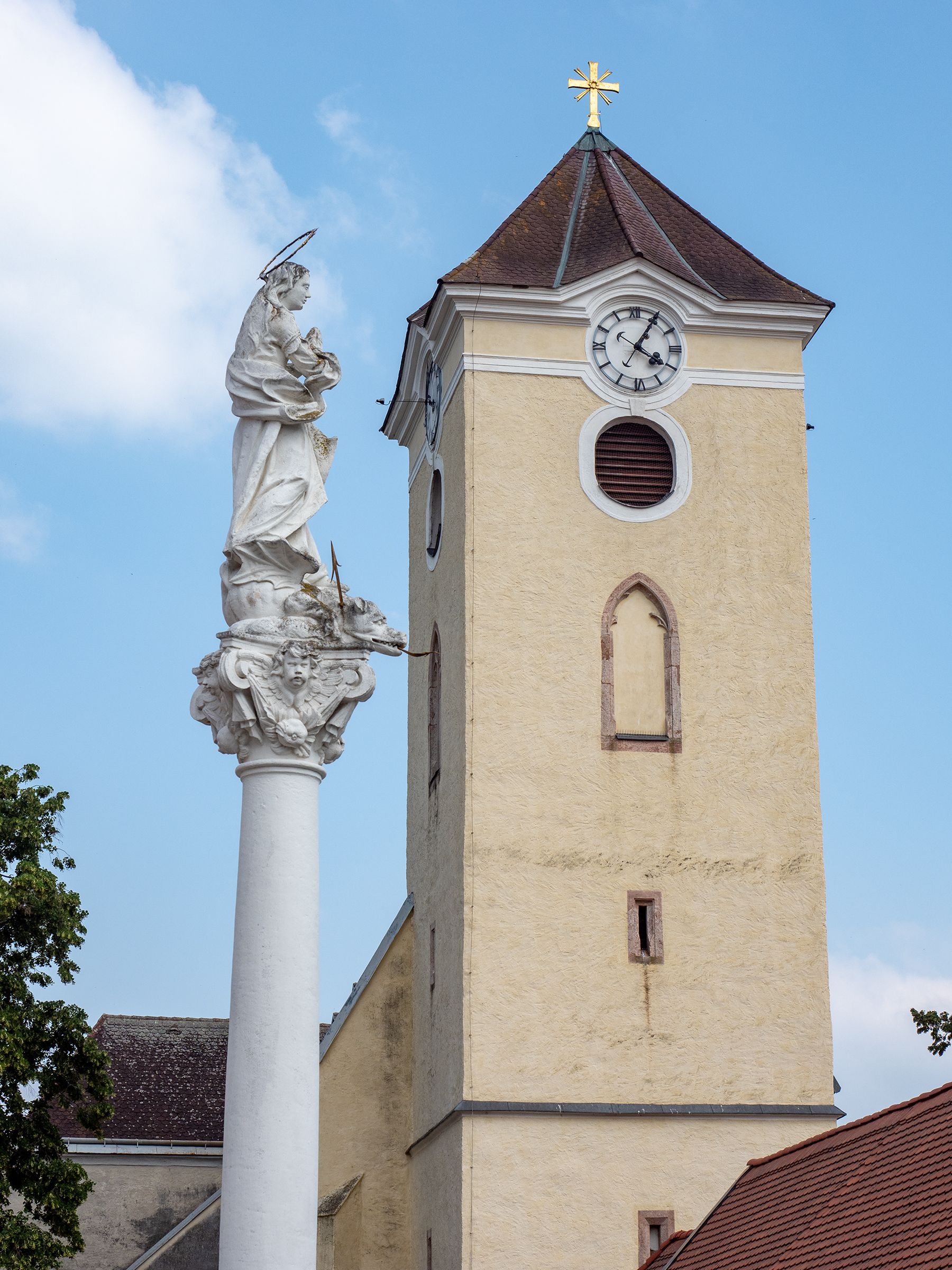 Kostolná veža s hodinami a mariánskym stĺpom v Schrattenthale na pozadí modrej oblohy.