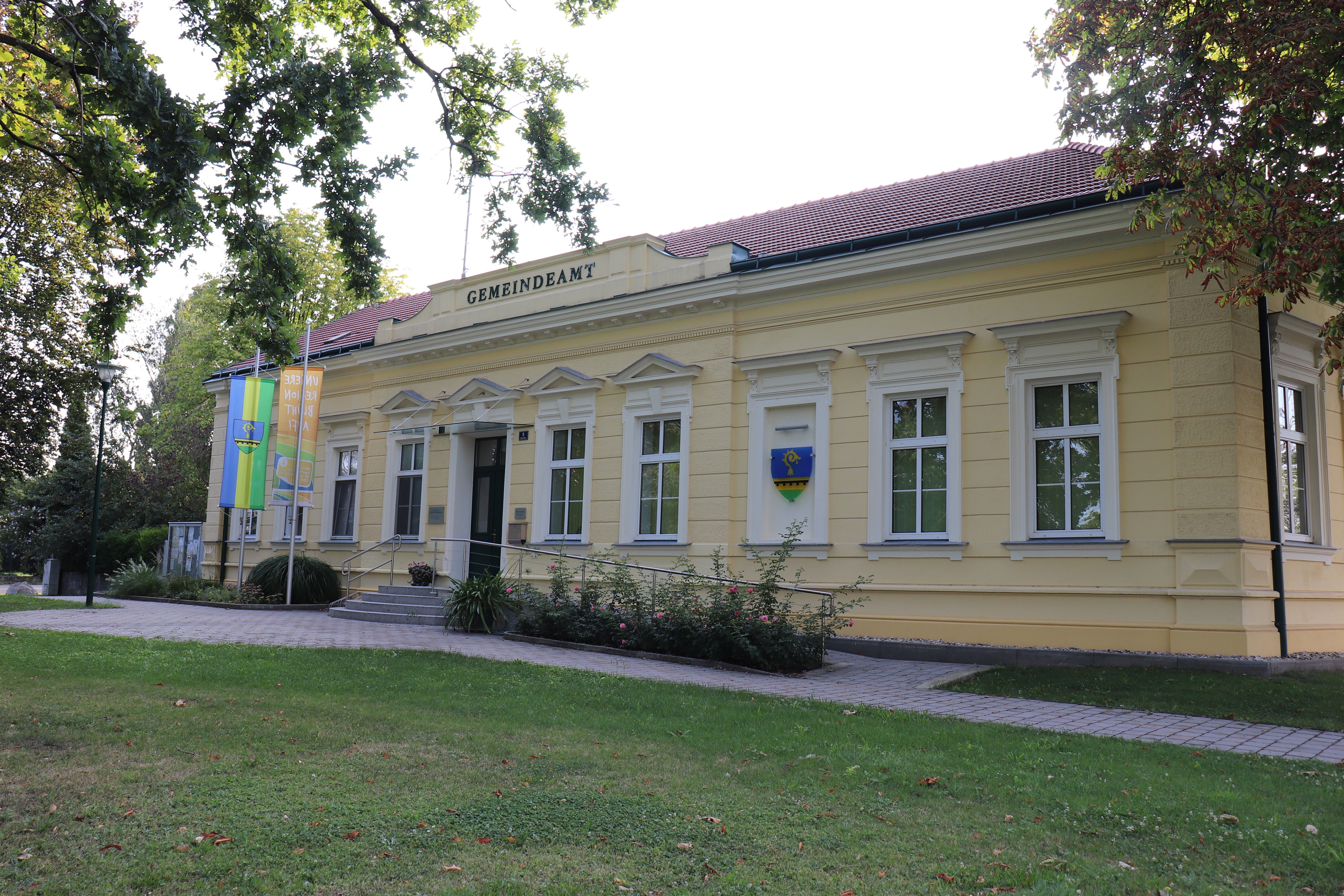Obecný úrad v Raasdorfe, svetlá administratívna budova s červenou strechou, vlajkami a erbom, obklopená stromami a trávnikom.