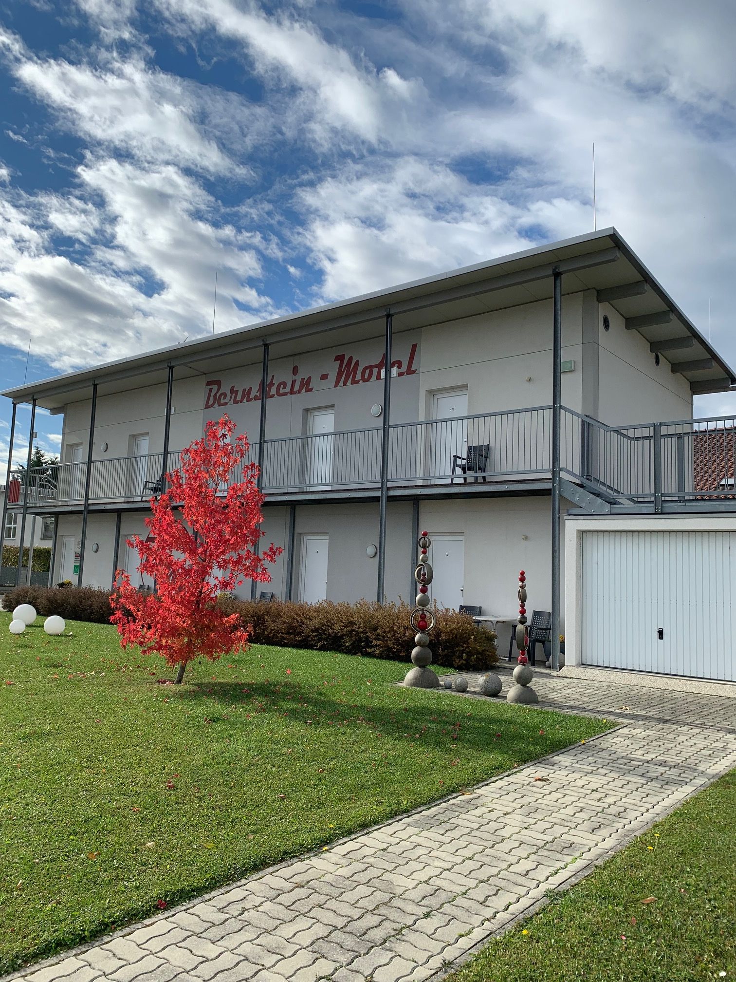 Dvojposchodová budova s nápisom "Bernstein Motel", červený strom v popredí, modrá obloha s mrakmi.