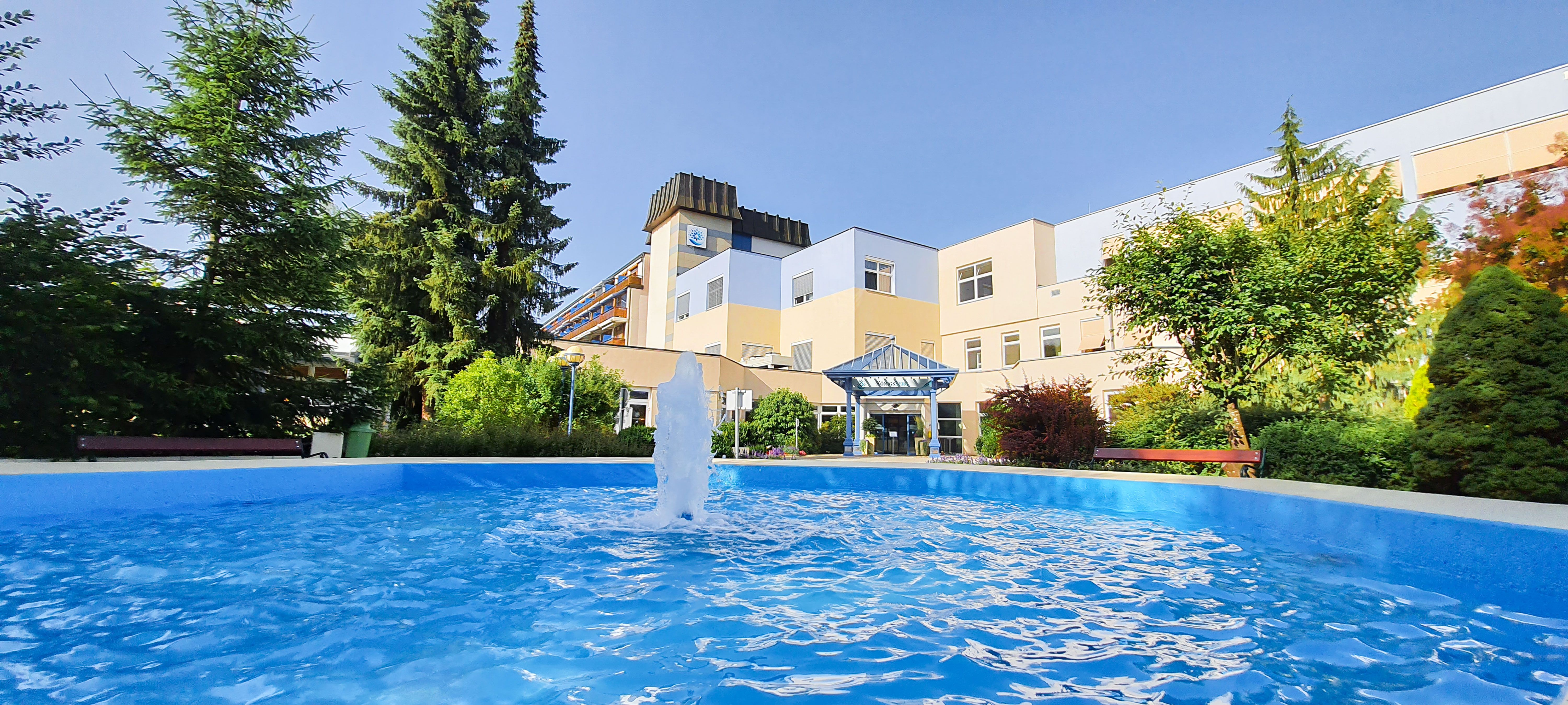 Exteriérový pohľad na kúpeľné stredisko s fontánou a záhradou.