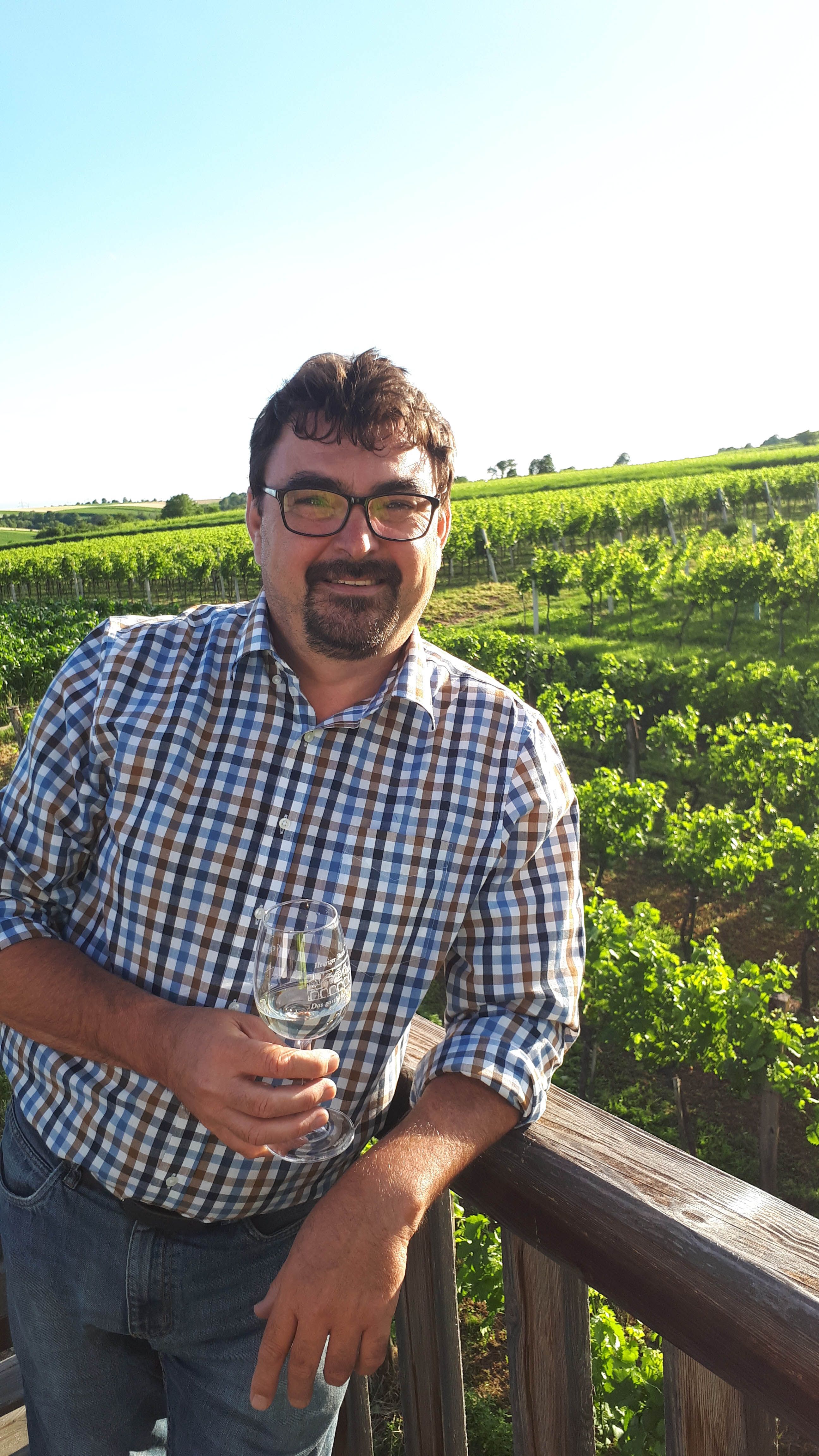 Muž v kockovanej košeli stojí pred vinicou s pohárom vína v ruke.