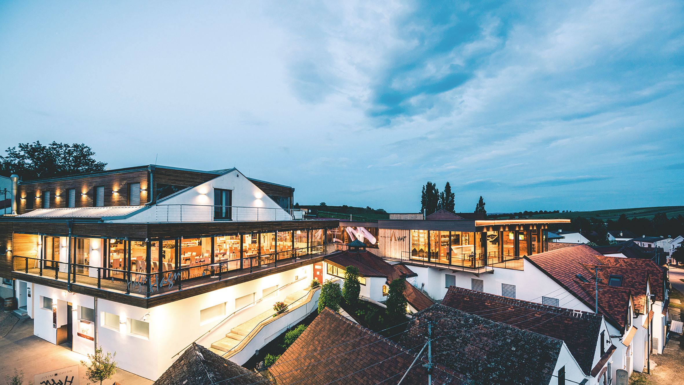 Moderná vinárska architektúra za súmraku s osvetlenými interiérmi a okolitými budovami.