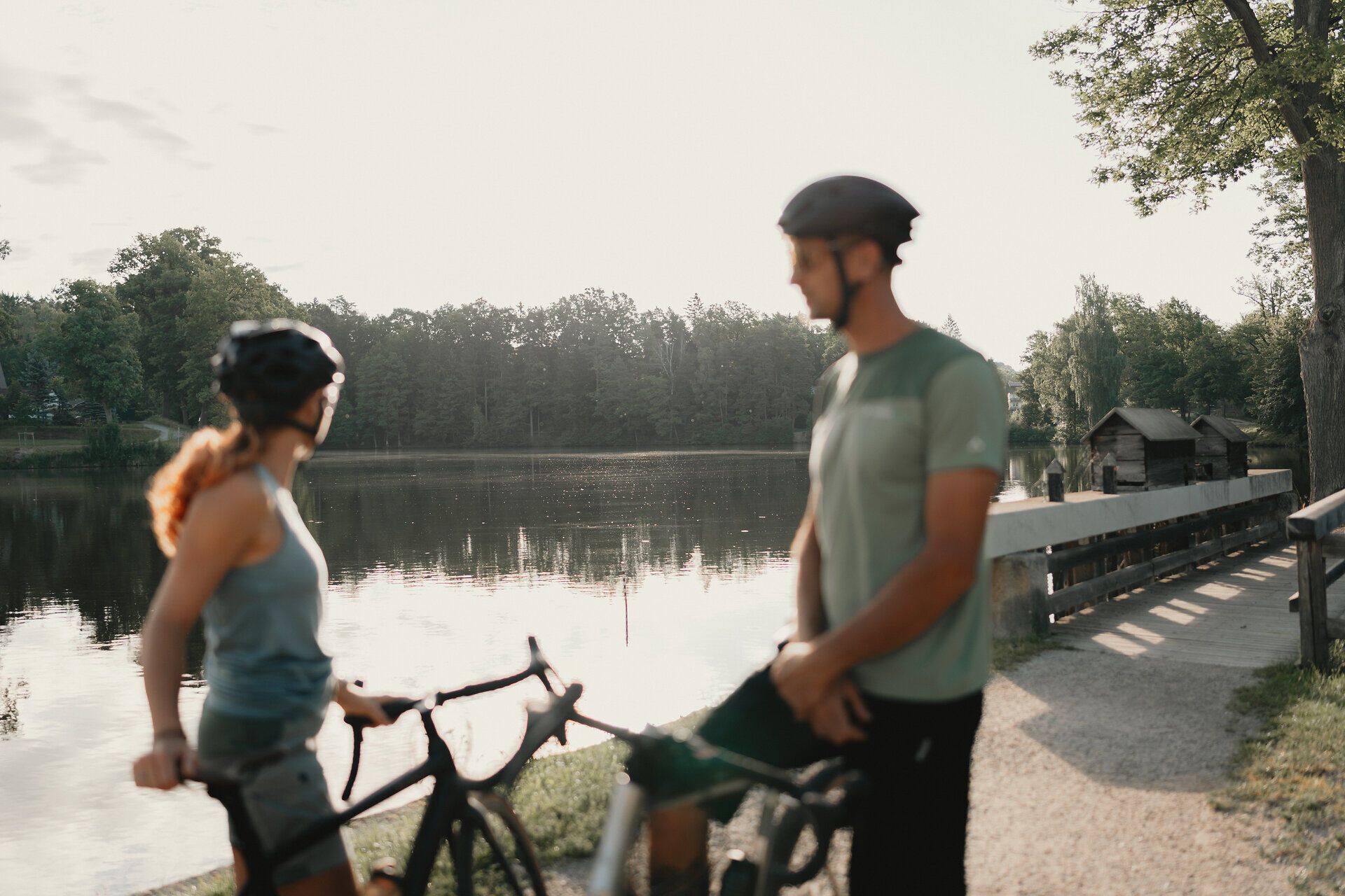 Dvaja cyklisti v prilbách stoja so svojimi bicyklami na brehu tichého rybníka obklopeného lesom.