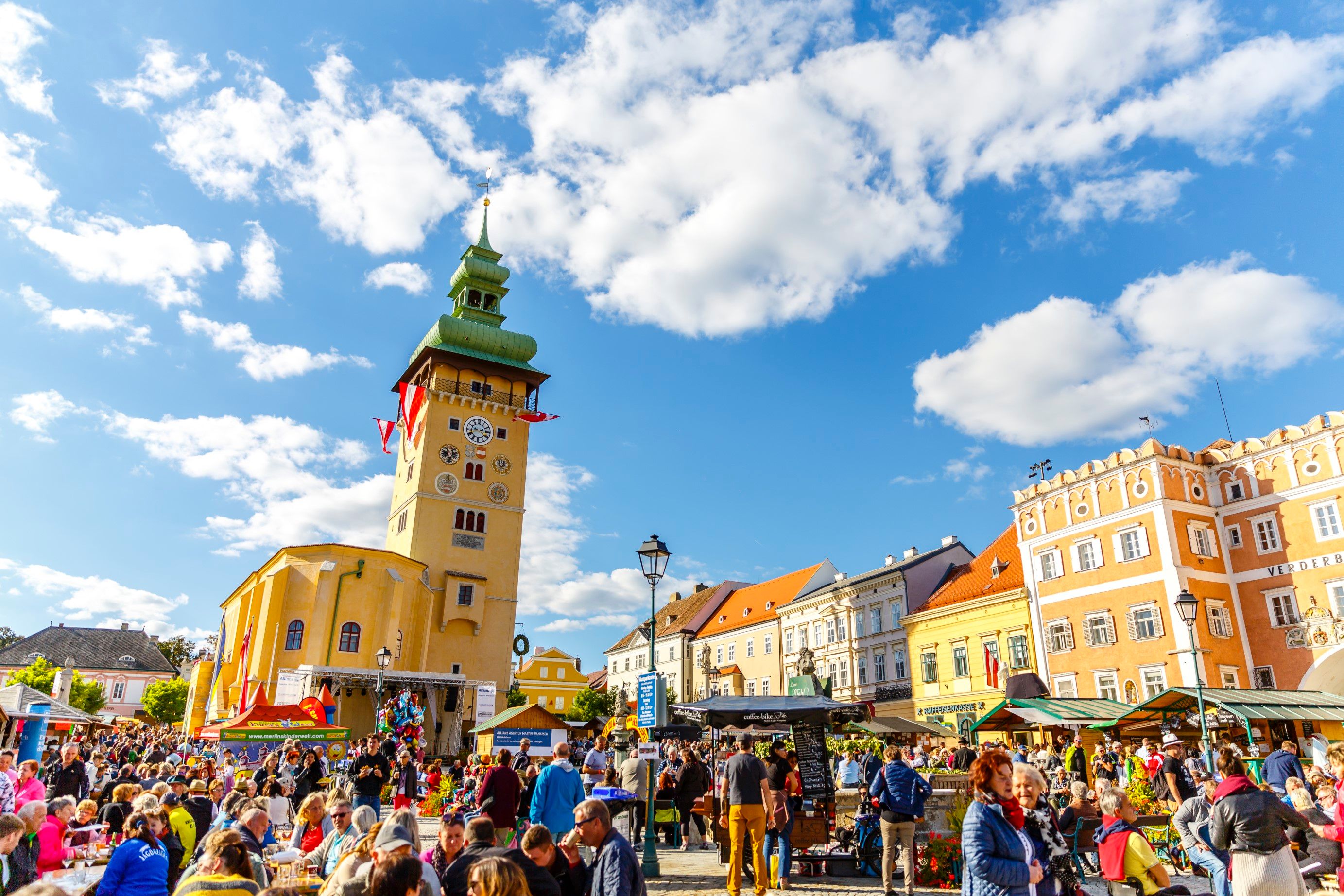 Dav ľudí na rušnom námestí v meste Retz s nápadnou hodinovou vežou v pozadí.