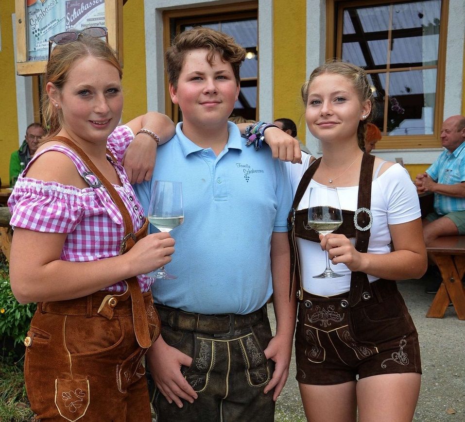Traja mladí ľudia v tradičnom bavorskom kroji s pohármi vína pred žltou budovou.