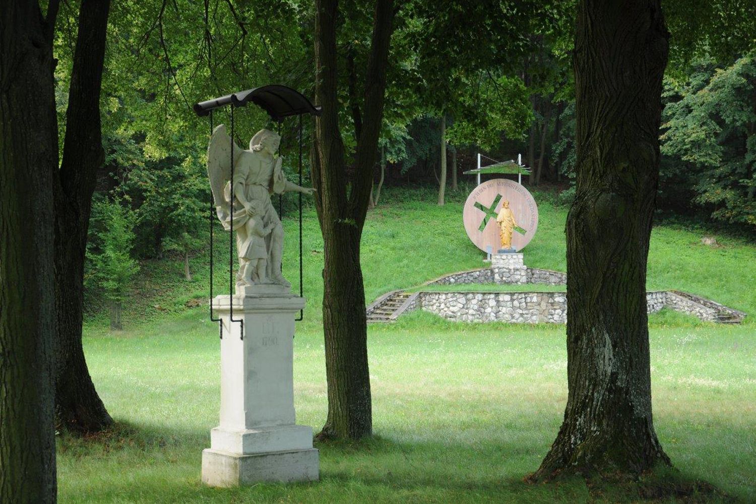 Socha anjela s dieťaťom v popredí, v pozadí ďalšia socha na podstavci v lesoparku.
