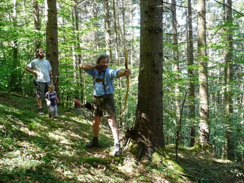 Osoba strieľa z luku v lese, v pozadí ďalšie dve osoby.