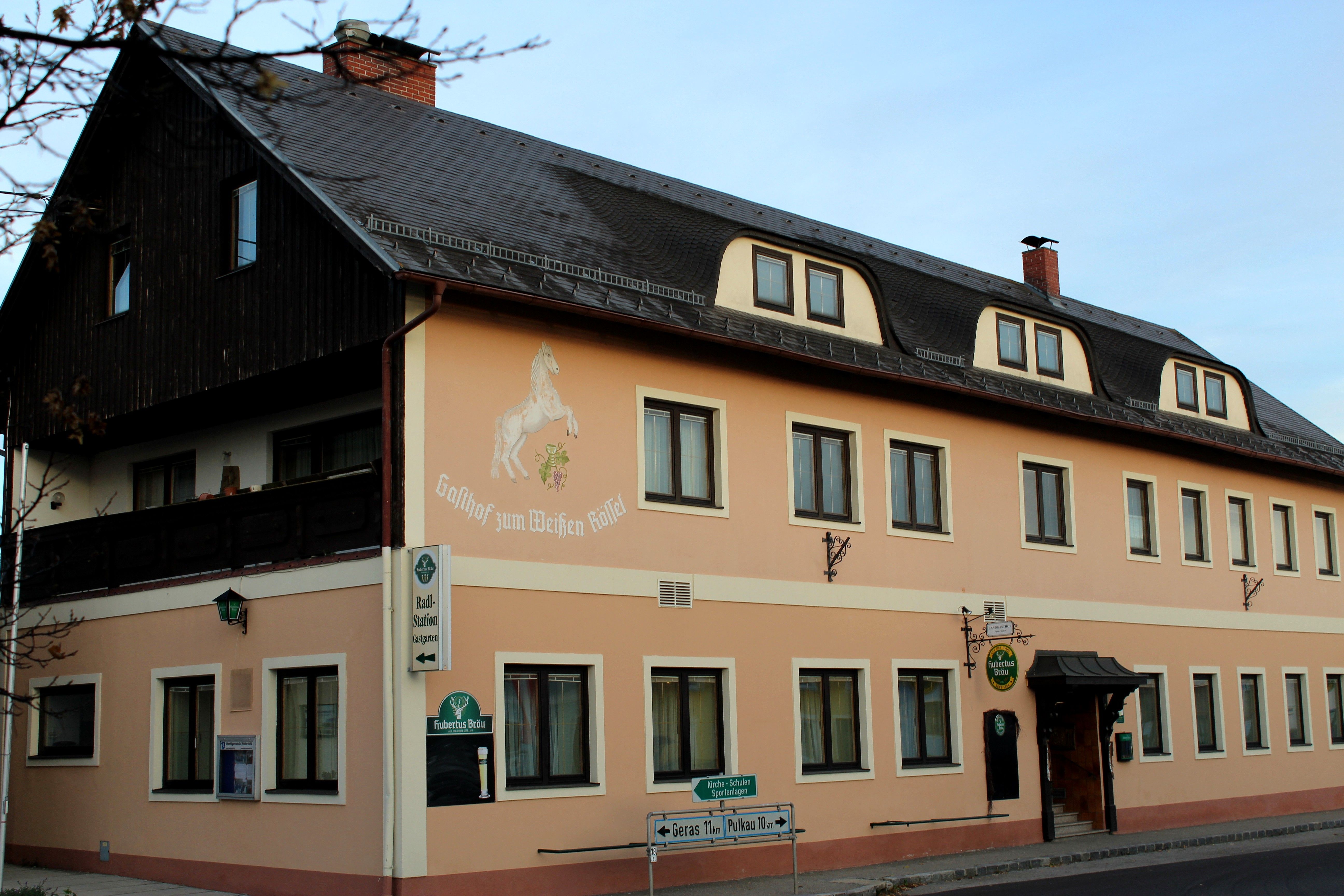 Exteriérový pohľad na hostinec "Zum weißen Rössel" s béžovou fasádou a maľbou koňa.