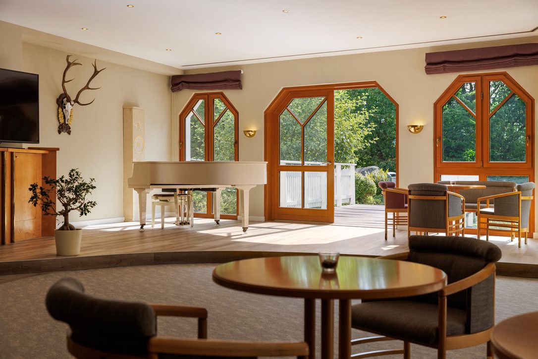 Svetlá izba s bielym klavírom, dreveným nábytkom a veľkými oknami, ktoré sa otvárajú na terasu s výhľadom na zeleň.