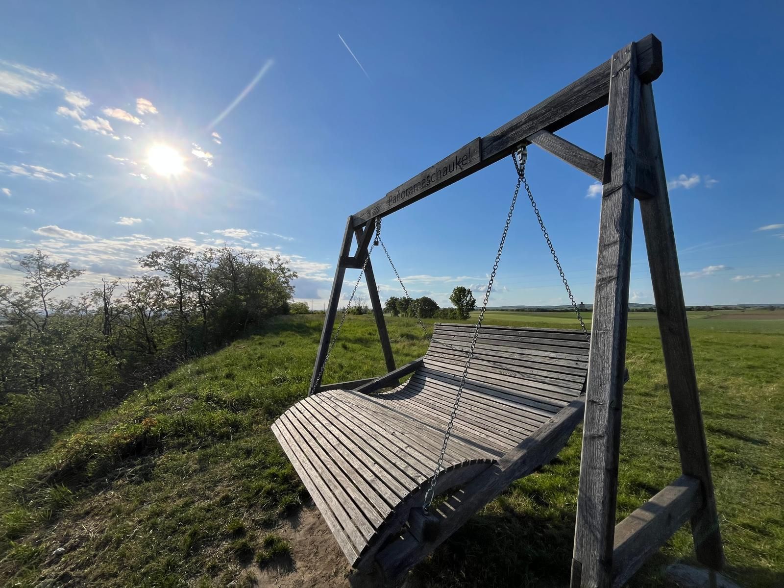 Veľká drevená panoramatická hojdačka na zelenej lúke pod jasne modrou oblohou. Nízke slnko osvetľuje krajinu a vrhá dlhé tiene, zatiaľ čo oblohu križujú jednotlivé mraky a parná stopa. Scéna pôsobí pokojne a prívetivo so širokým výhľadom na polia a prírodu.