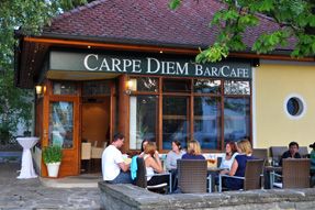 Pohľad zvonku na kaviareň Carpe Diem, ľudia sedia pri vonkajších stoloch.