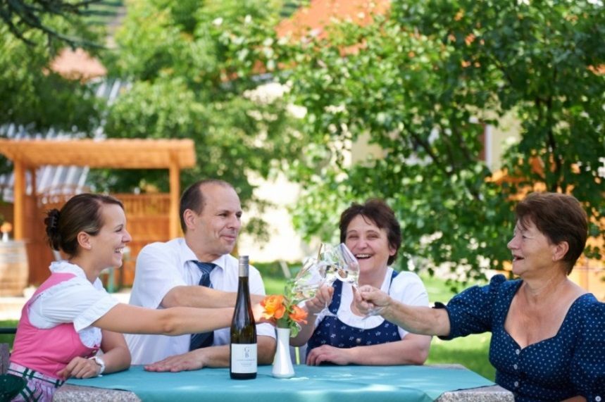 Štyria ľudia si pri stole vonku cinknú pohárom vína.