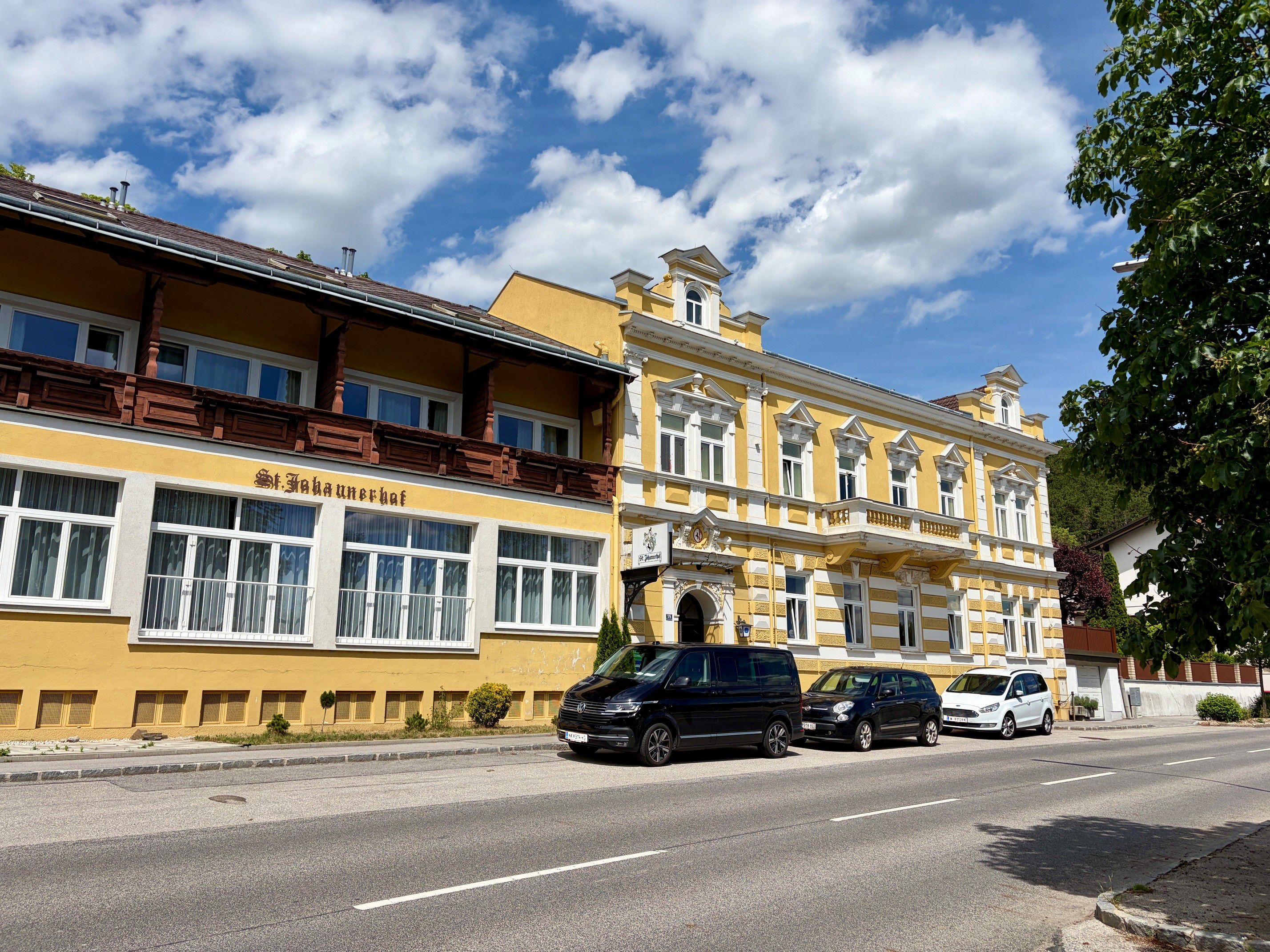 Žltá budova s nápisom "St. Johannerhof" na ceste, obklopená zaparkovanými autami a stromami.