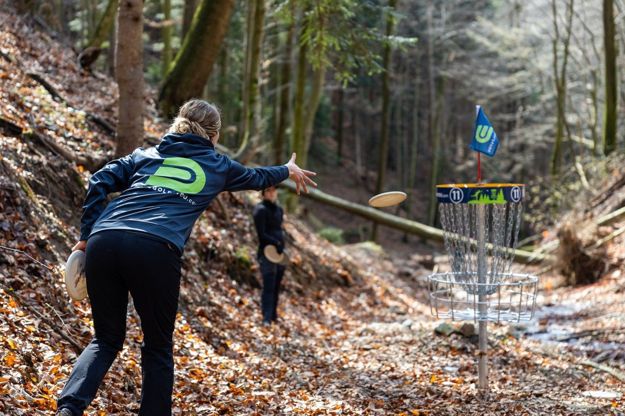 Osoba hádže frisbee do discgolfového koša v lese.