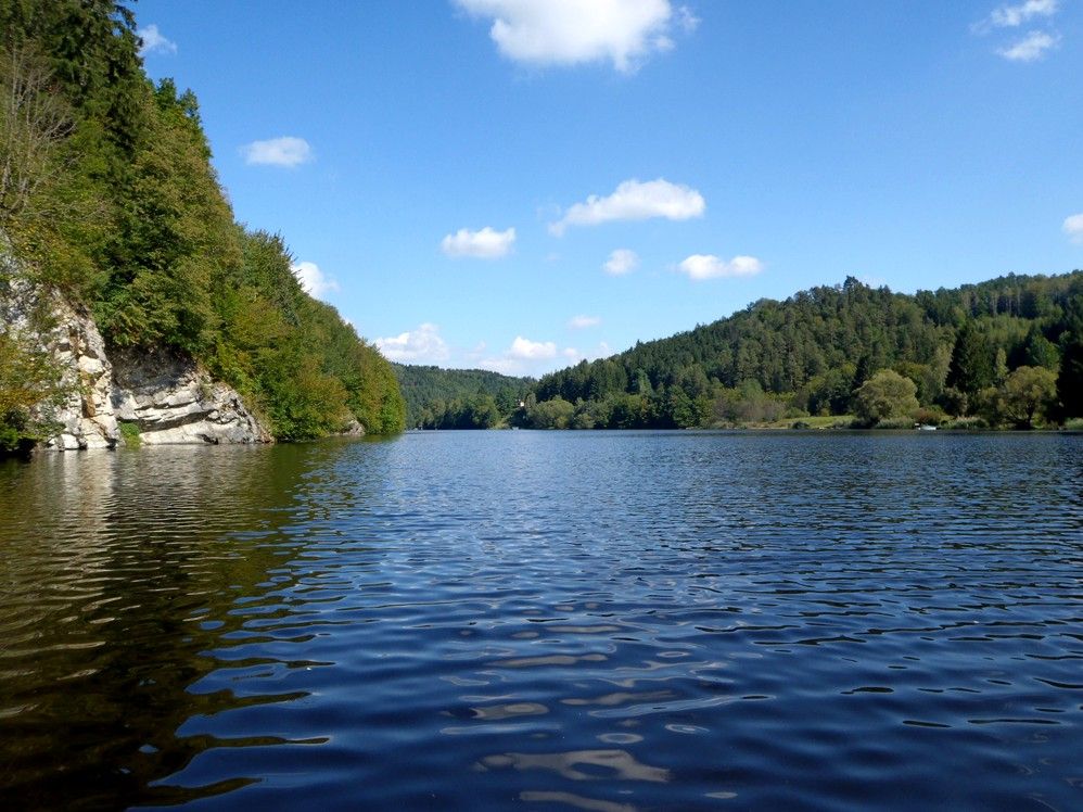 Pokojné jazero obklopené zalesnenými kopcami pod modrou oblohou s niekoľkými mrakmi.