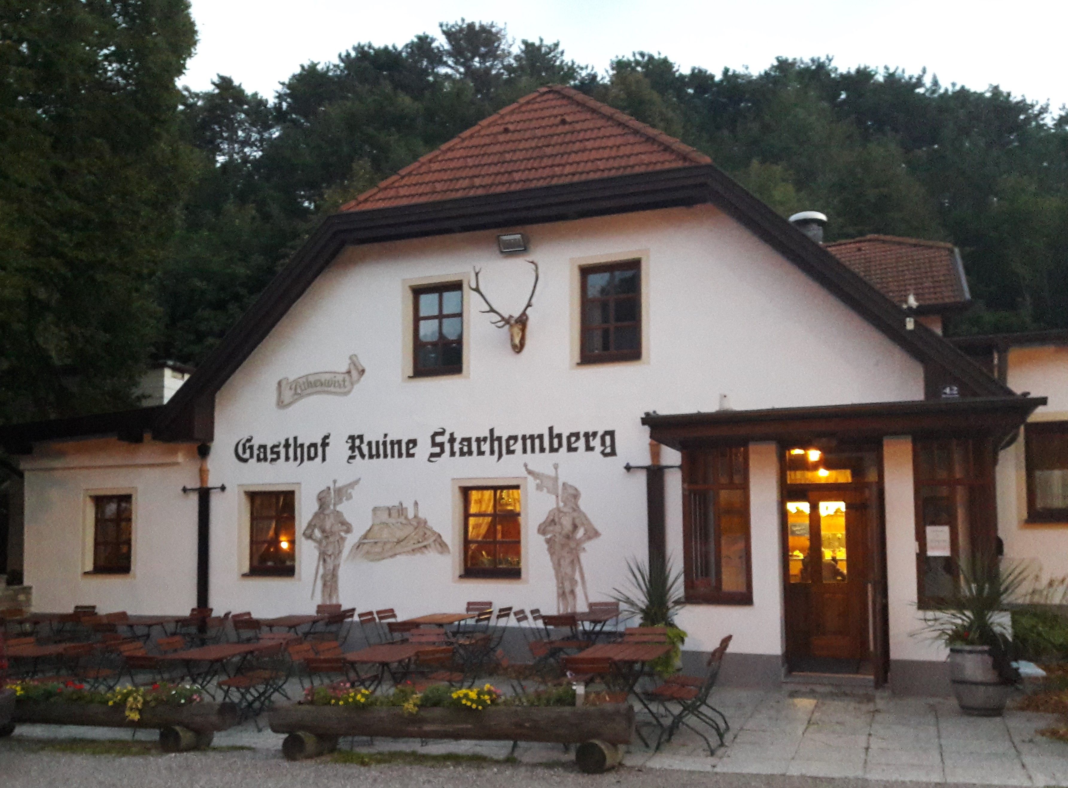 Tradičný hostinec s vonkajším priestorom a nástennými maľbami, obklopený stromami.