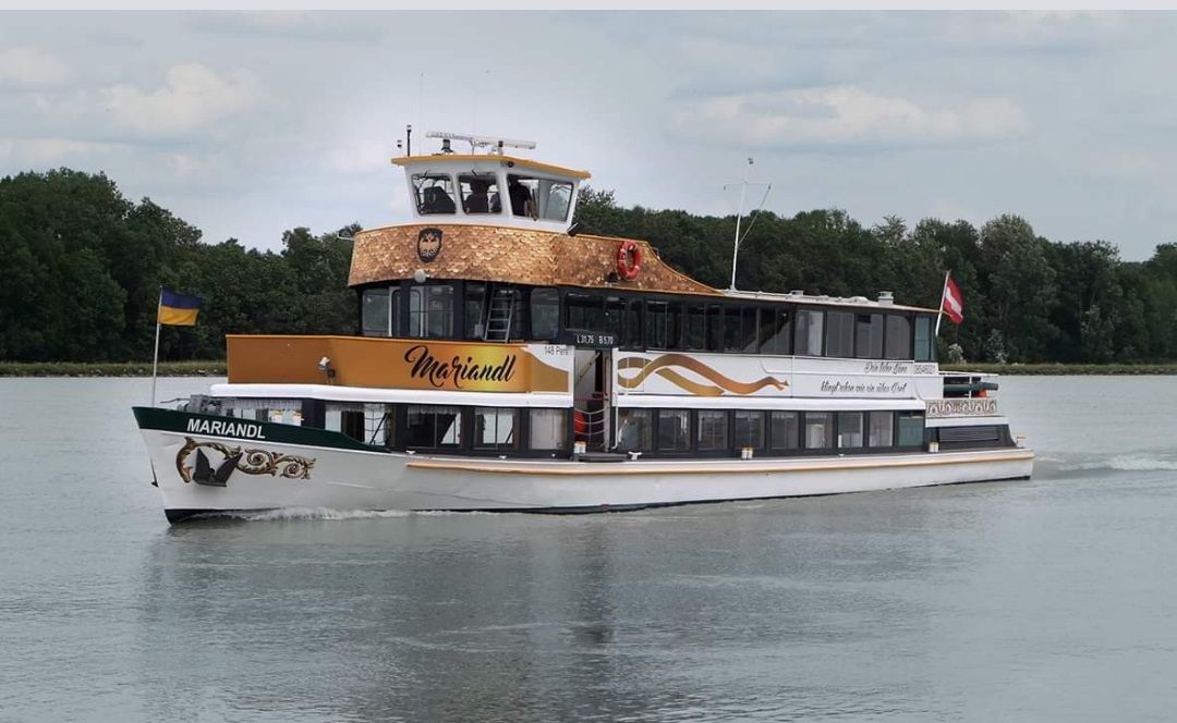 Výletná loď MS Mariandl sa plaví po rieke obklopenej stromami. Nesie vlajky Ukrajiny a Rakúska.