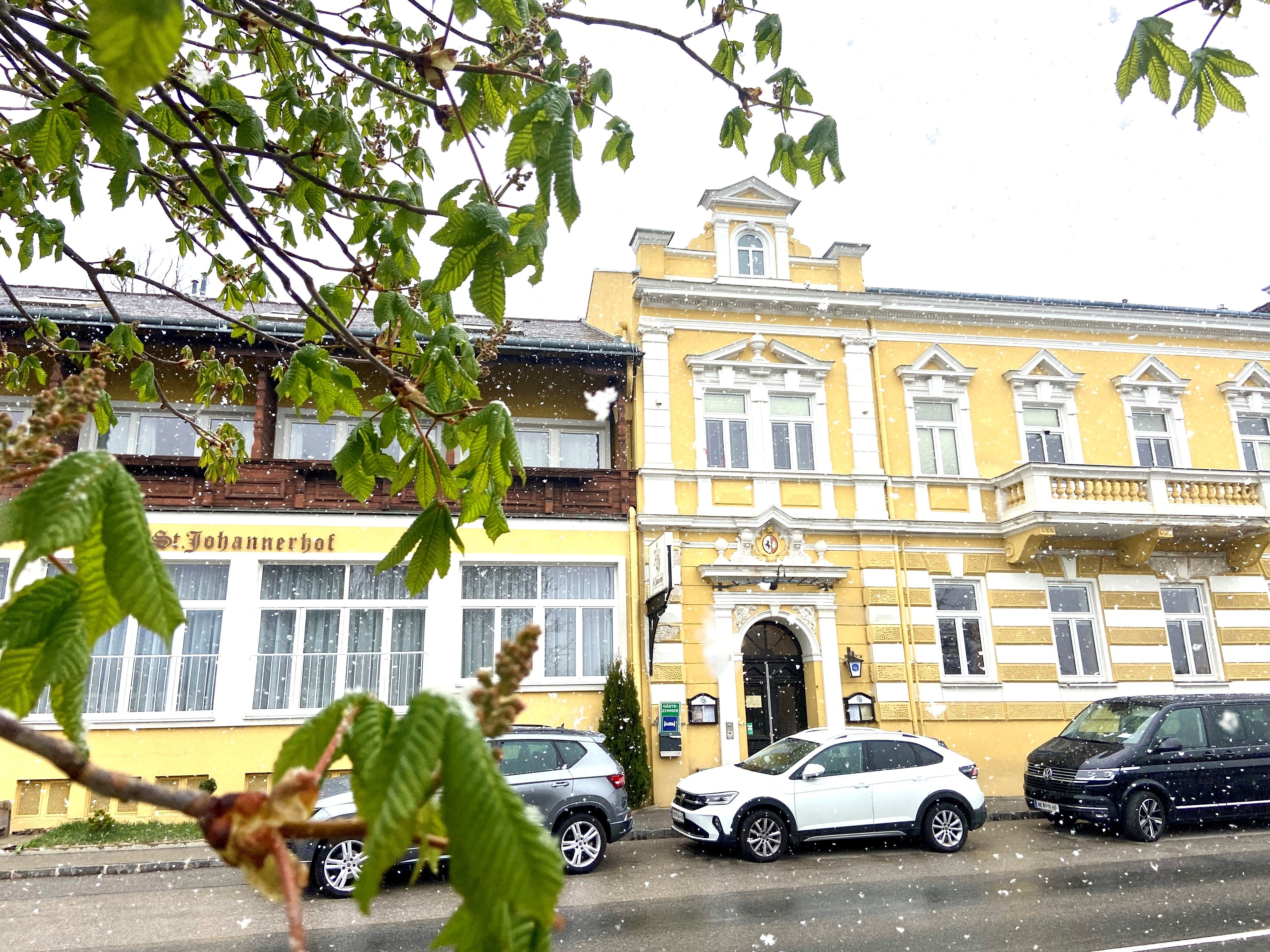 Žltá budova Sankt Johannerhof so zasneženými autami pred ňou a zeleným lístím v popredí.