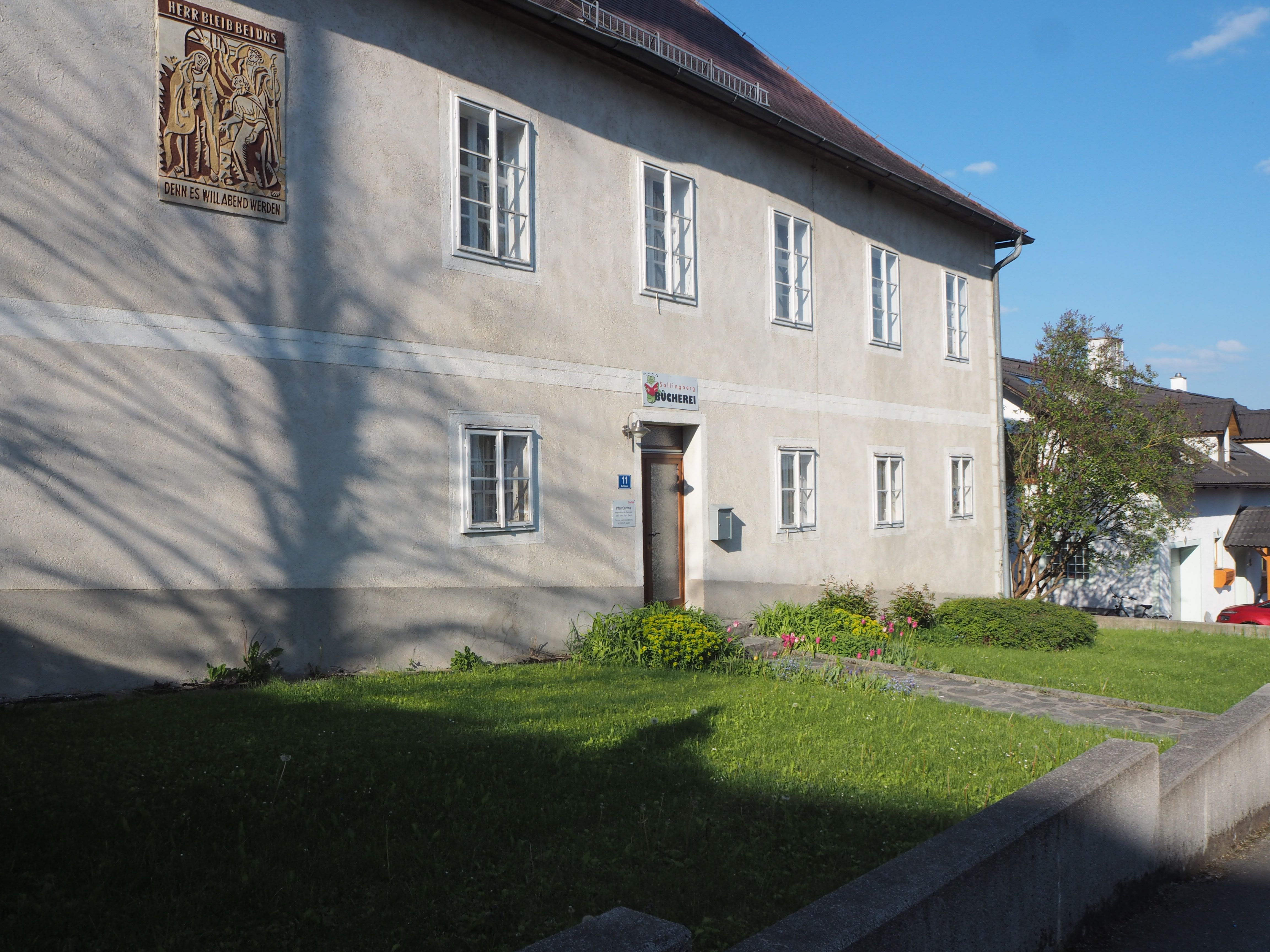 Dvojposchodová budova s niekoľkými oknami a nápisom nad dverami, obklopená udržiavanou záhradou.
