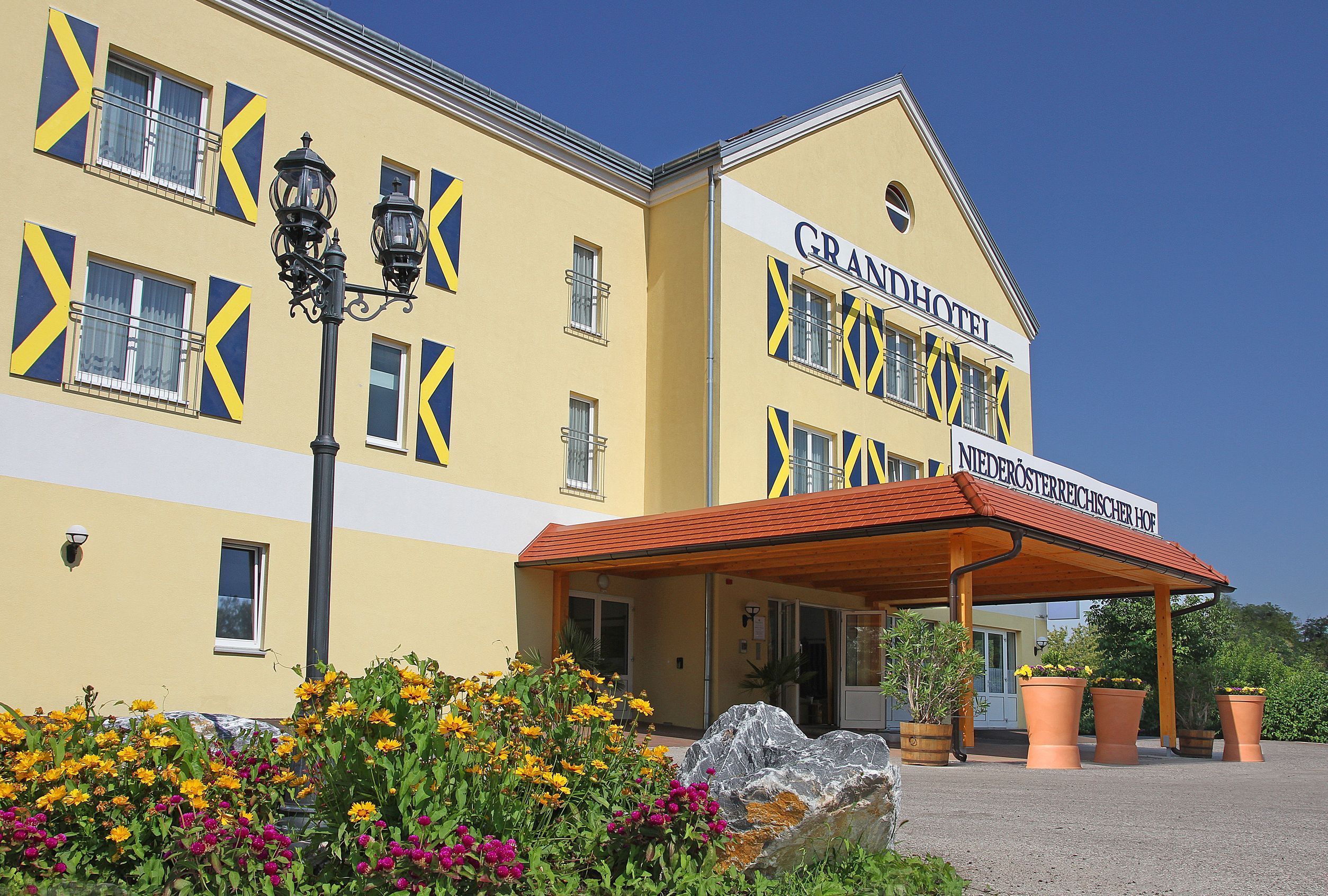Exteriérový pohľad na žltý hotel s modrými okenicami a baldachýnom. Pred budovou kvitnú žlté a ružové kvety.