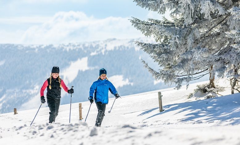 Dvaja ľudia na bežkách pri bežkovaní na zasneženej trase s horami v pozadí.
