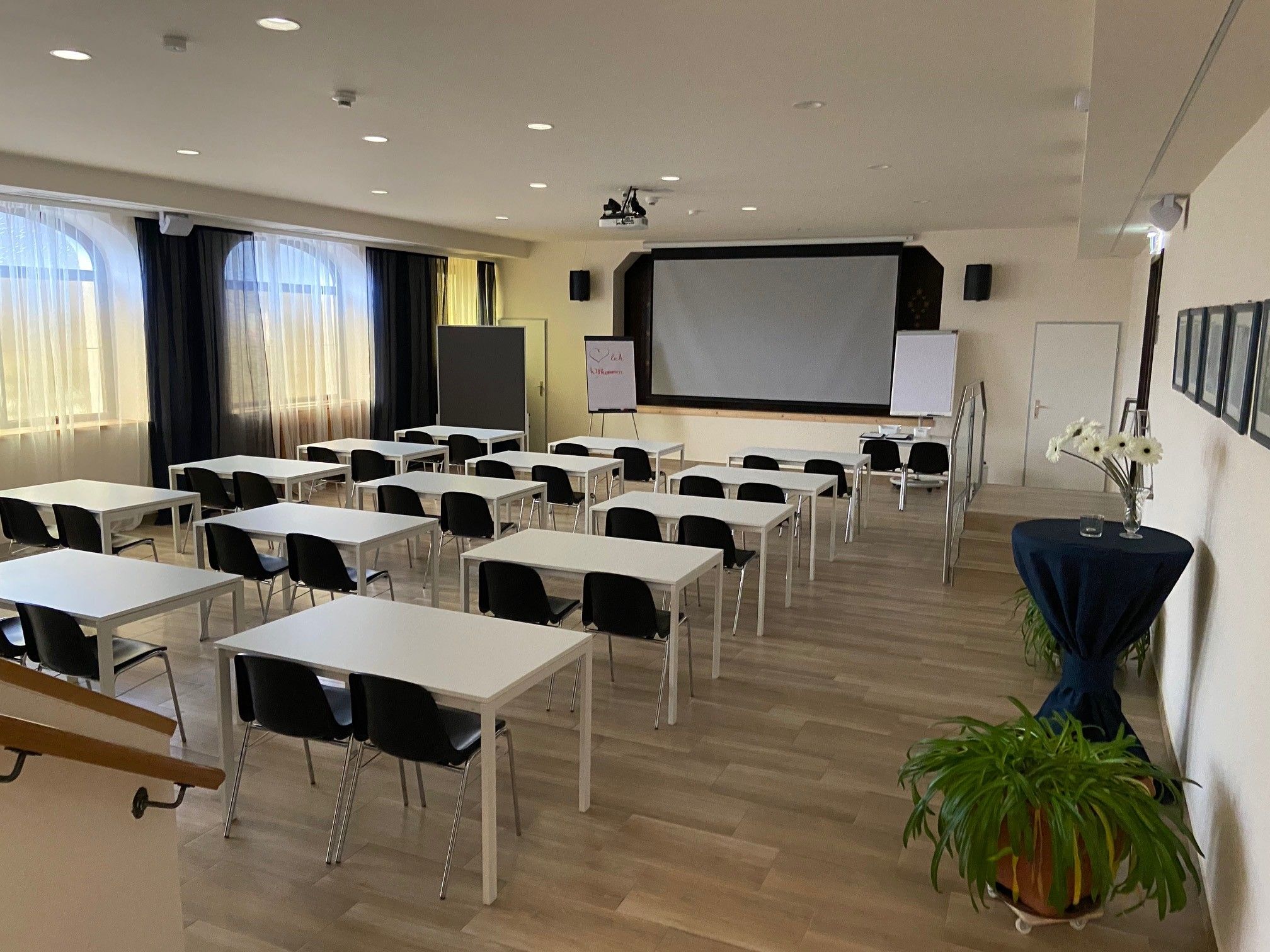 Prázdna seminárna miestnosť s bielymi stolmi, čiernymi stoličkami a veľkou obrazovkou vpredu.
