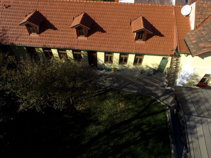 Letecký pohľad na budovu s červenou škridlovou strechou a niekoľkými oknami, obklopenú záhradou.