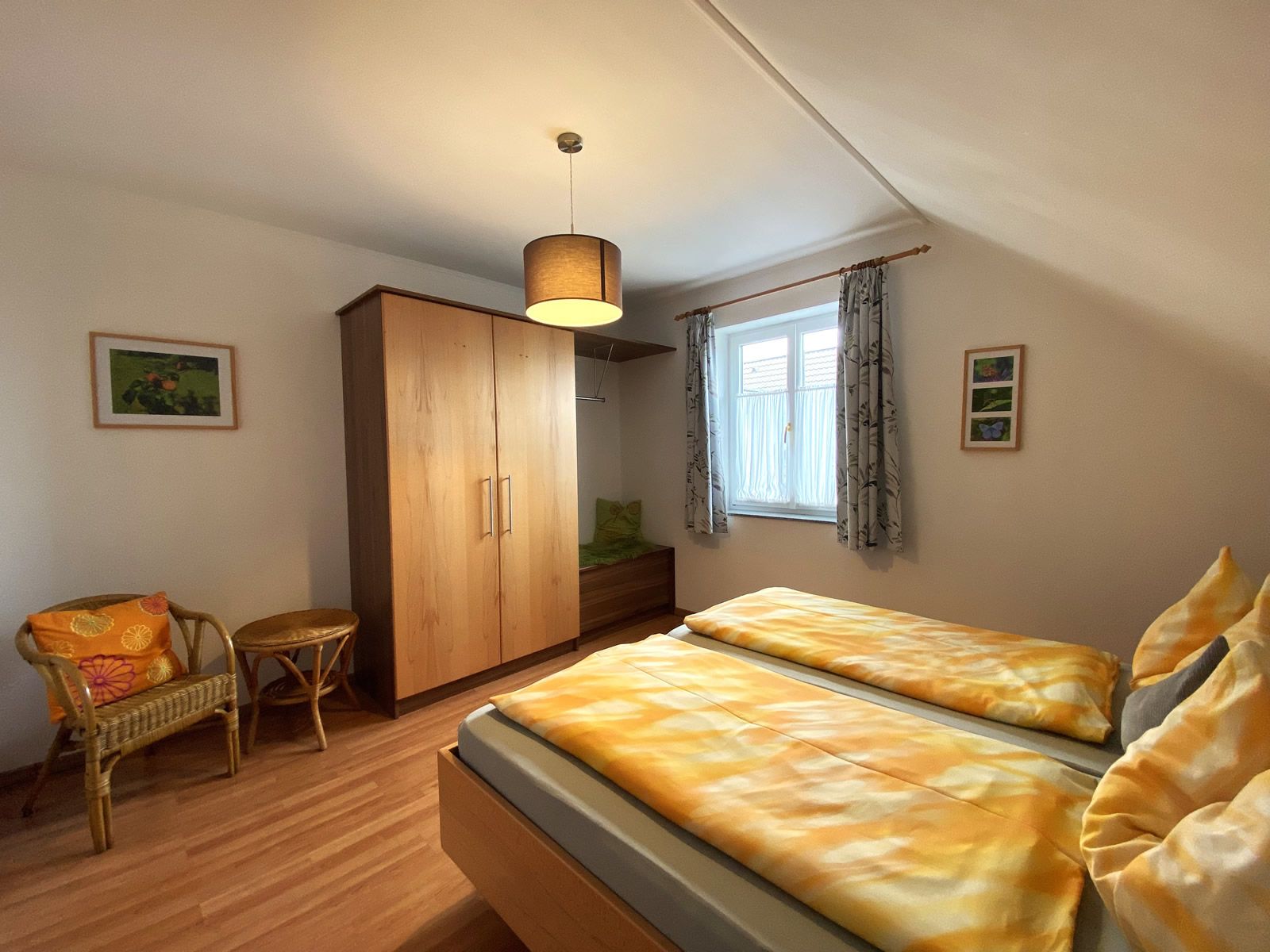 Spálňa s manželskou posteľou, drevenou skriňou, prúteným kreslom a oknom so závesmi.