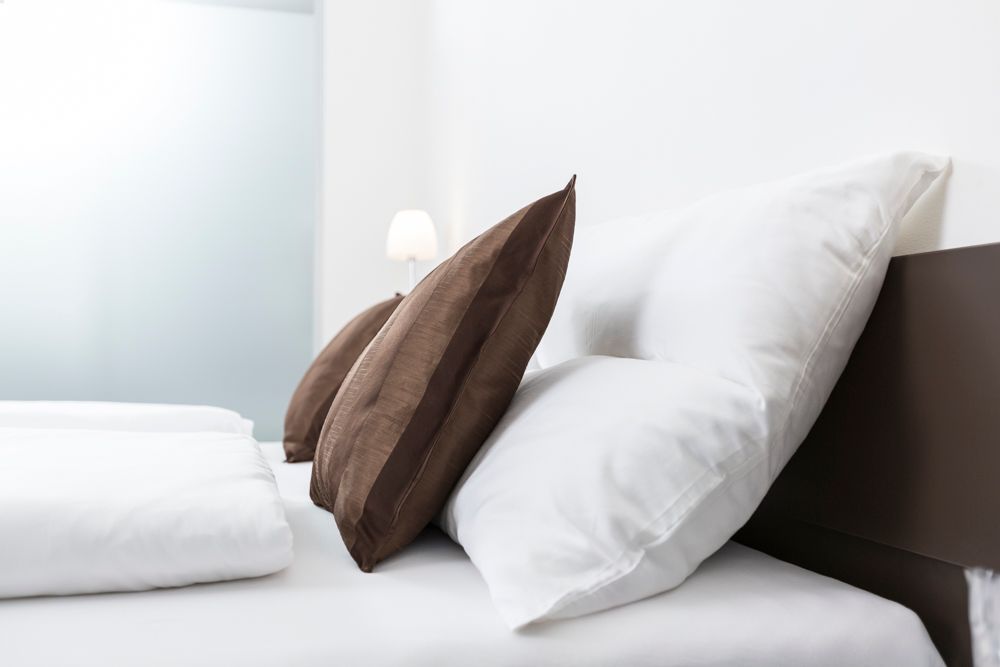 Posteľ s bielymi vankúšmi a hnedými ozdobnými vankúšmi, lampa v pozadí.