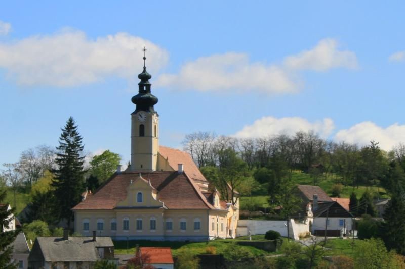 Kostol s vežou vo vidieckom prostredí, obklopený stromami a domami.