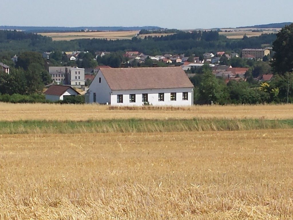 Biely dom s červenou strechou stojí na poli, v pozadí je mesto a les.