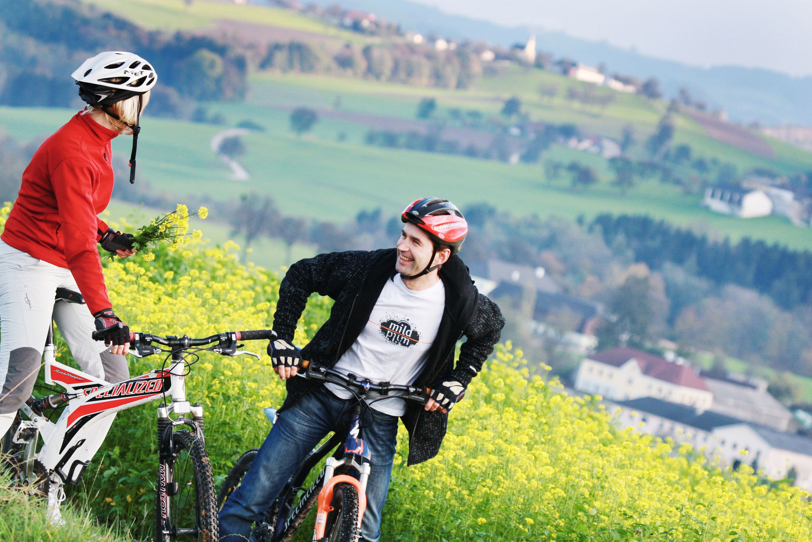 Dvaja cyklisti s prilbami stoja na rozkvitnutom poli so zelenou krajinou v pozadí.