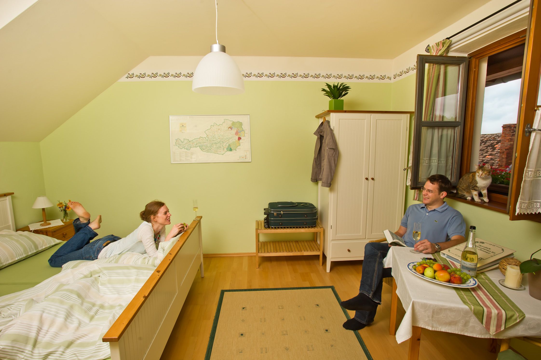 Útulná izba pre hostí s relaxačným párom. Žena leží na posteli, muž sedí za stolom. Pri okne sedí mačka.