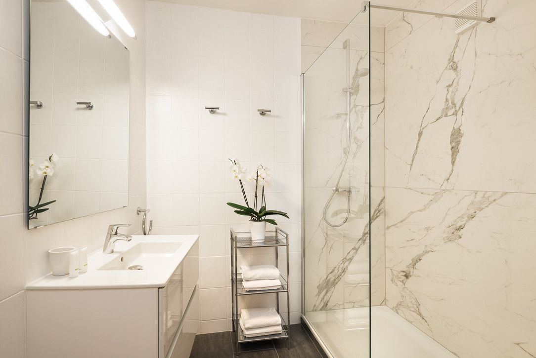 Moderná kúpeľňa s bielym umývadlom, zrkadlom, sklenenou sprchou a orchideou na poličke.