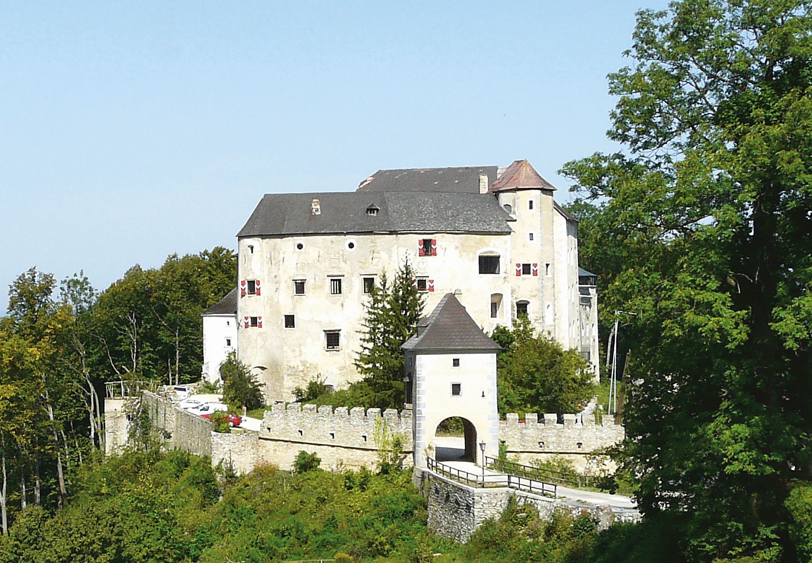 Historický hrad s bielou fasádou a bojovými hradbami, obklopený stromami.