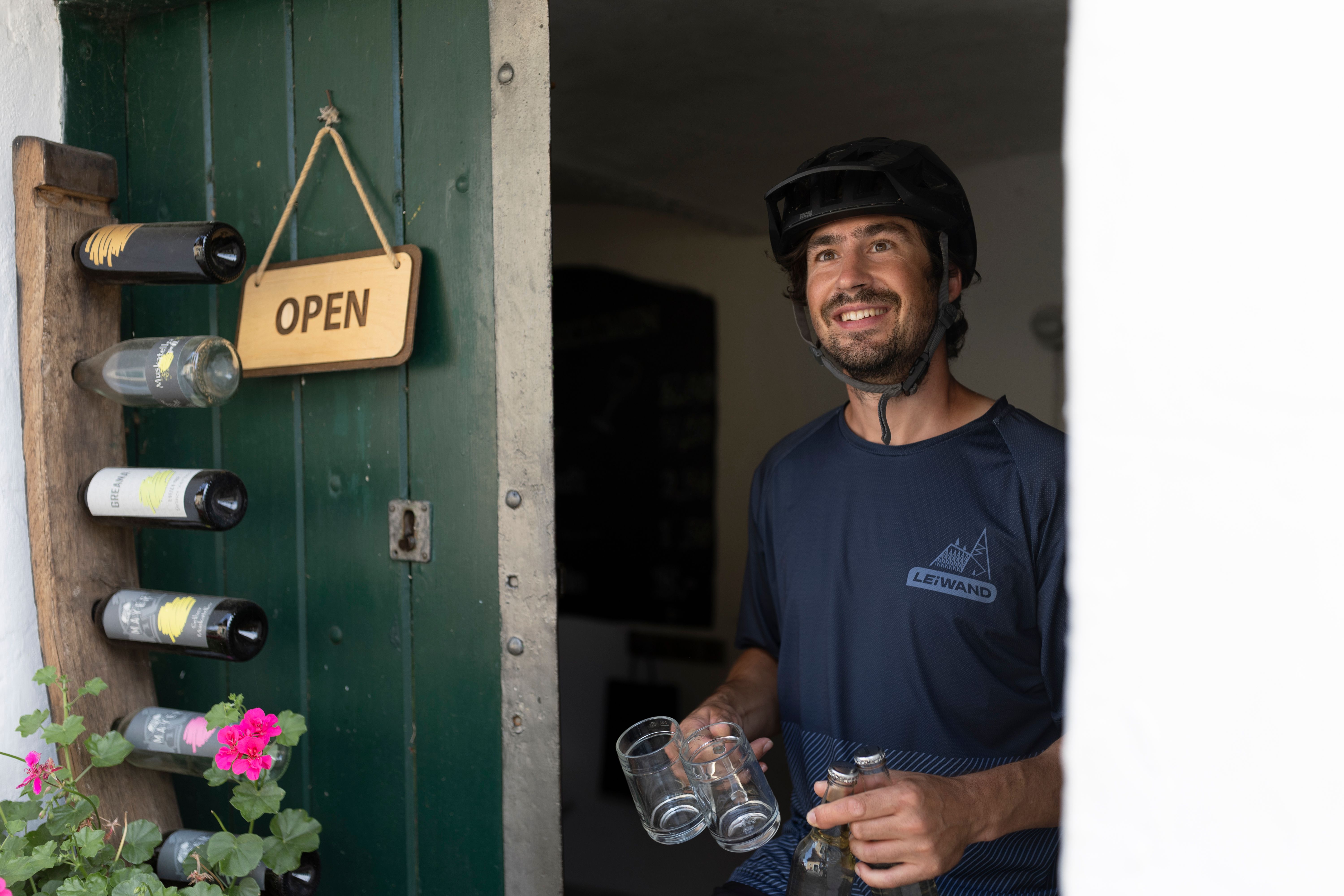 Muž s cyklistickou prilbou stojí s úsmevom v otvorených dverách, v rukách drží okuliare a fľaše. Na dverách visí nápis "Otvorené".