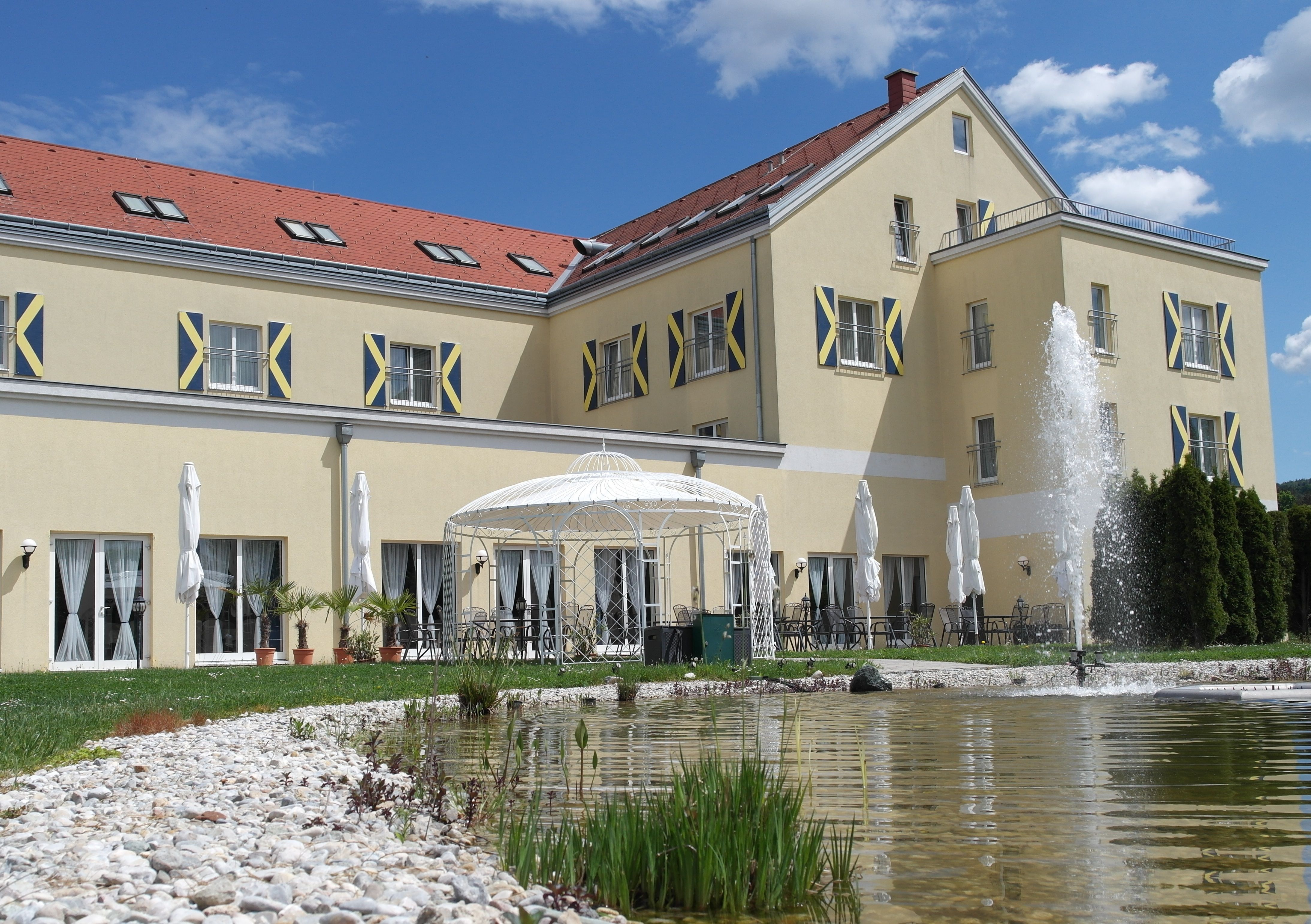 Veľká žltá budova s červenými škridlami, fontánou a pavilónom v popredí.