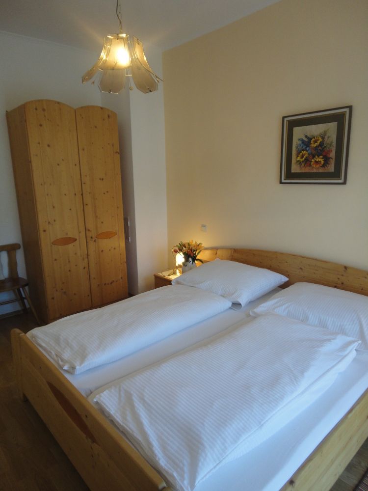 Útulná spálňa s manželskou posteľou, drevenou skriňou, nočným stolíkom s kvetmi a obrazom na stene.