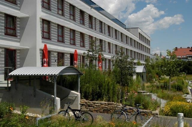 Moderná budova s niekoľkými poschodiami, obklopená zelenou krajinou a bicyklami v popredí.