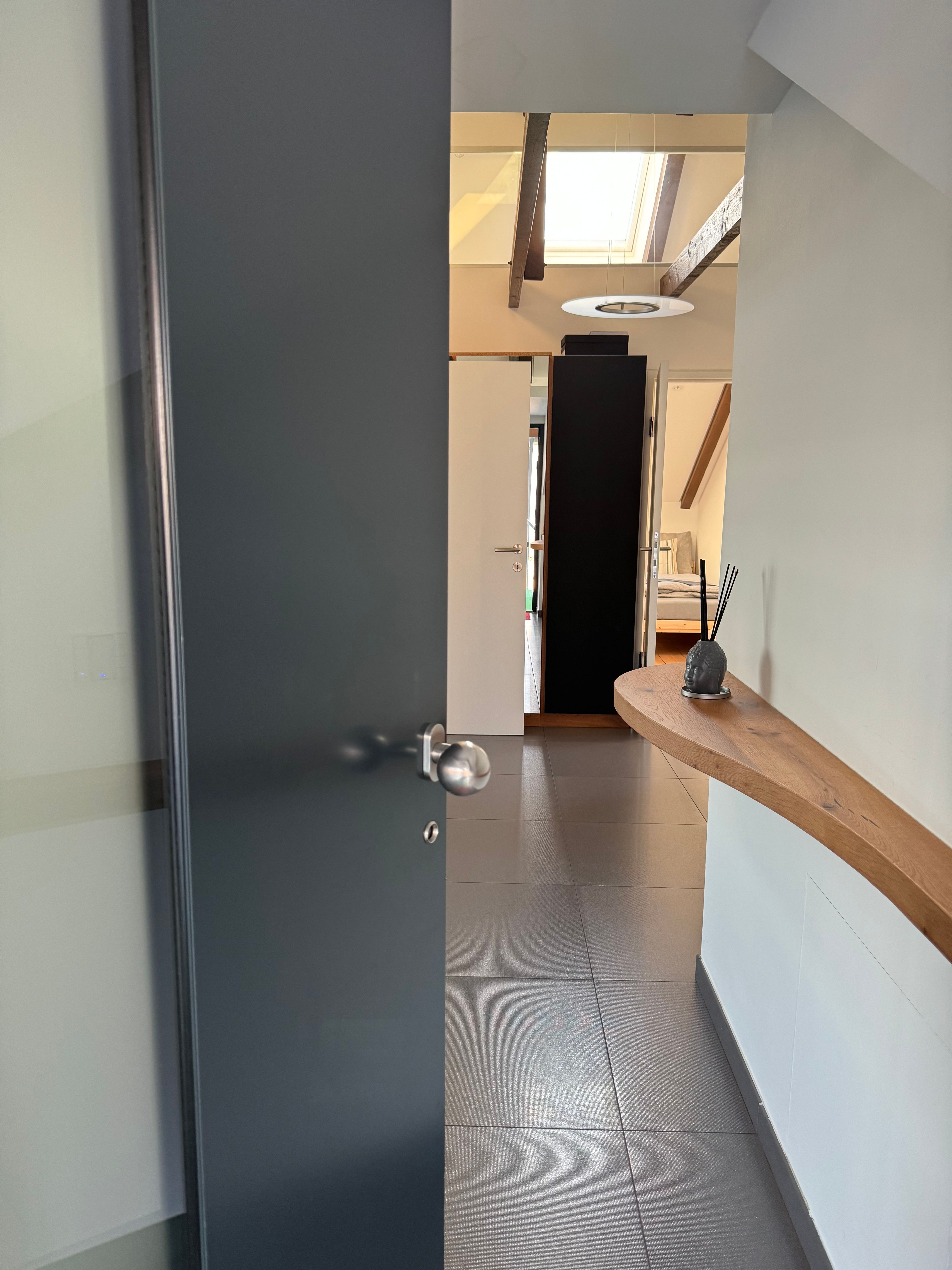 Moderný vstupný priestor s otvorenými dverami, dláždenou podlahou a drevenou policou na stene.